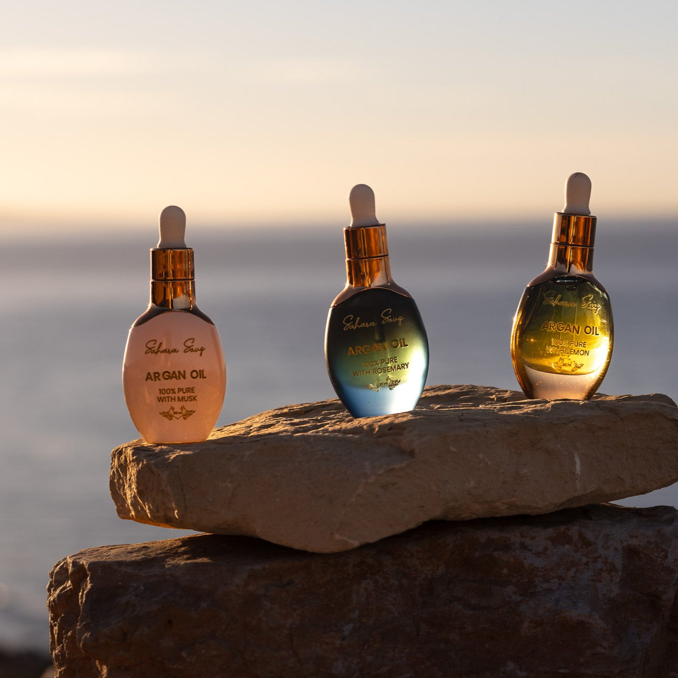 Three bottles of Sahara Souq's Moroccan Argan Oil Bundle Set with different essential oil blends placed on a rock by the sea, each bottle representing a different blend: Argan Oil with Lemon Essential Oil, Argan Oil with Rosemary Essential Oil, and Argan Oil with Musk. Three bottles of Sahara Souq's Moroccan Argan Oil Bundle Set with different essential oil blends placed on a rock by the sea, each bottle representing a different blend: Argan Oil with Lemon Essential Oil, Argan Oil with Rosemary Essential Oil, and Argan Oil with Musk.