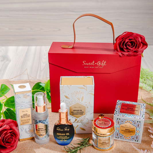 A gift set containing skincare products, including a jar of rose water, a bottle of argan oil with rosemary, and a clay mask pack, all branded with Sahara Souq, arranged in front of a red box with 'Sweet Gift' text.